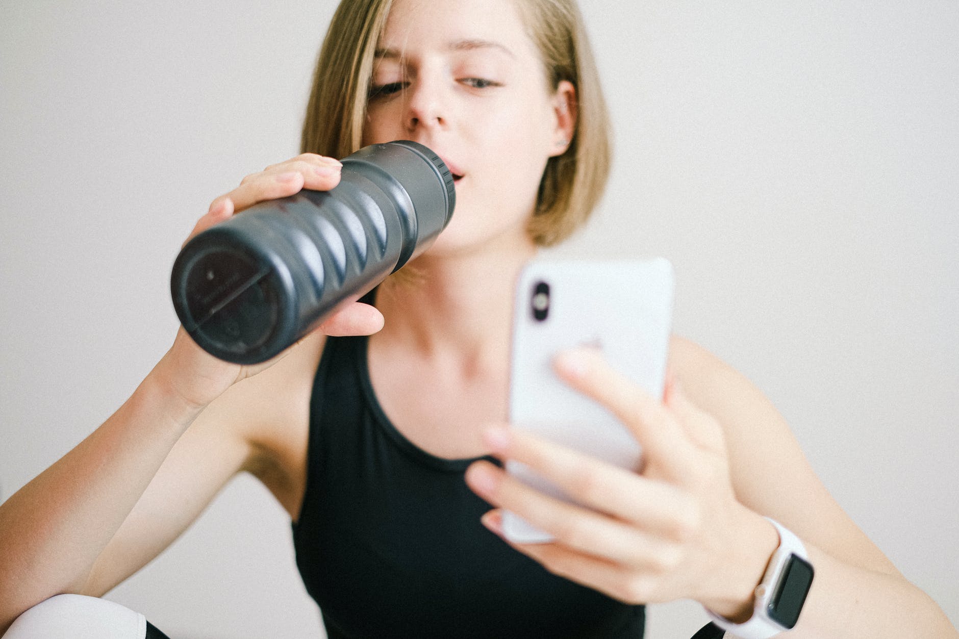 woman in black tank top holding white smartphone while drinking
